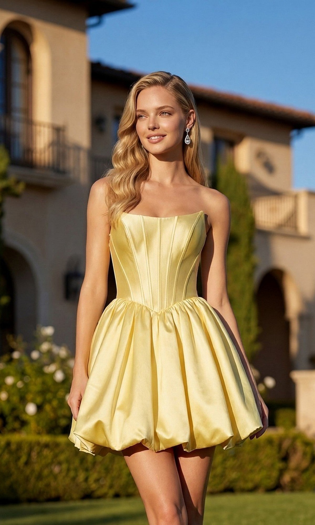 A woman wears the Juliet JT7184U Short Homecoming Dress in yellow, featuring a strapless fitted corset bodice and voluminous A-line skirt, as she stands outdoors in front of a Mediterranean-style building.